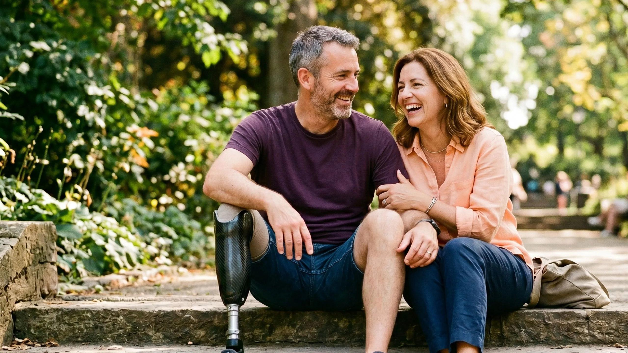 A couple sitting close together on sunny park steps, laughing; one has a below-knee prosthetic leg